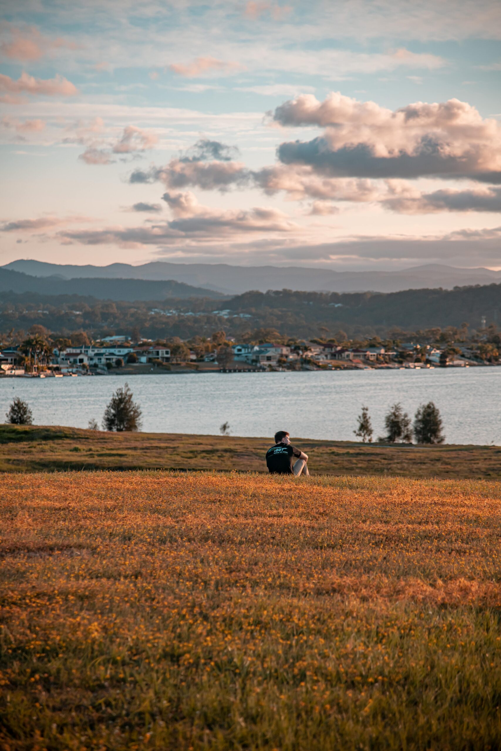 Serene scene of a man sitting by a lake at sunset in Robina, QLD, with mountains and homes in view.